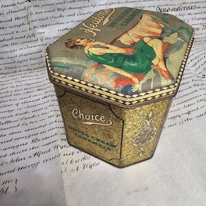 May include: Vintage Heaton's Swiss Cream Caramels tin. The tin has a hexagonal shape with a decorative lid featuring a woman in a green dress. The tin is gold with the word "Choice" and "Swiss Cream Caramels" on the front.