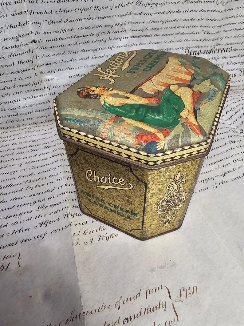 May include: Vintage Heaton's Swiss Cream Caramels tin. The tin has a hexagonal shape with a decorative lid featuring a woman in a green dress. The tin is gold with the word "Choice" and "Swiss Cream Caramels" on the front.