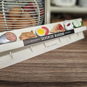 Puede incluir: Un juego vintage de marcadores para sándwiches con réplicas de alimentos en miniatura, como salami, pollo, huevo, tomate, jamón y pepino. Los marcadores están adheridos a un soporte de cartón blanco con el texto "SANDWICH MARKERS" y "MADE IN ENGLAND".