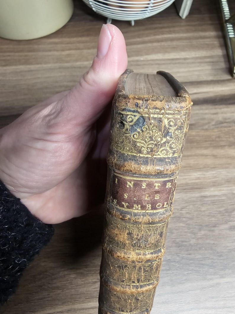 K&ouml;nnte beinhalten: Antikes Buch mit einem abgenutzten, braunen Ledereinband. Der Buchr&uuml;cken weist goldgepr&auml;gte florale Muster und ein rotes Etikett mit dem Titel "INSTR SYMBOL" auf. Die Kanten des Buches weisen Alters- und Abnutzungserscheinungen auf.
