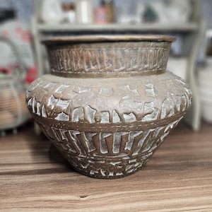 May include: A copper-colored, antique-style vase with intricate embossed patterns. The vase has a wide, rounded body and a slightly flared rim. The embossed design features repeating geometric and animal motifs, adding a touch of vintage charm to the piece.