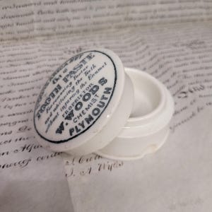 May include: A small, white ceramic jar with a lid. The lid is printed with black text that reads "Tooth Paste" and "W. Woods Chemist Plymouth". The jar is open, revealing its interior.