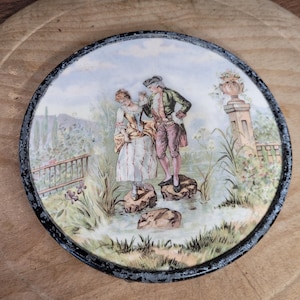 May include: Round decorative plate with a painted scene of a couple in a garden setting. The plate features a black rim and a detailed illustration with pastel colors, depicting a man and woman standing on rocks near water.