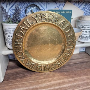 以下が含まれることがあります： 縁に「Give Us This Day Our Daily Bread」という文字が刻まれた、金色の金属製のプレート。プレートはハンマーで叩いたような質感です。