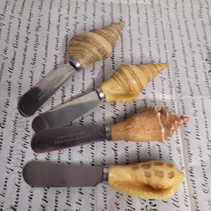 May include: Set of four seashell-shaped butter knives. The knives are made of stainless steel and have wooden handles carved in the shape of seashells. The handles are a light brown color and have a natural wood grain.