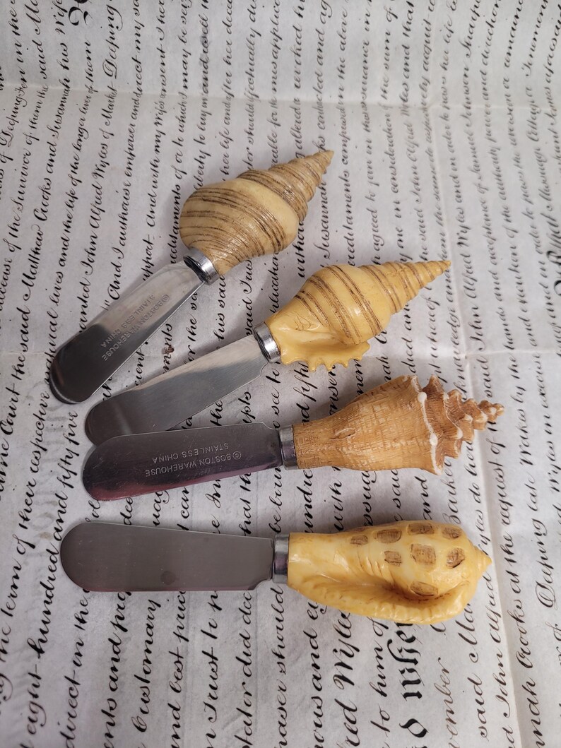May include: Set of four seashell-shaped butter knives. The knives are made of stainless steel and have wooden handles carved in the shape of seashells. The handles are a light brown color and have a natural wood grain.
