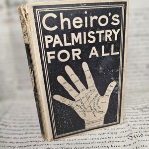 May include: An antique book, "Cheiro's Palmistry For All", with a black cover and a hand diagram. The cover shows wear and tear, indicating its age. The title is in white, and the book is placed on a background of old paper.