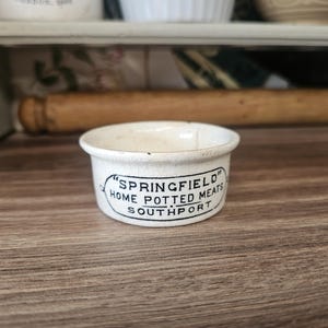 May include: Small, off-white ceramic bowl with the text "SPRINGFIELD HOME POTTED MEATS SOUTHPORT" in black. The bowl is round with a slightly flared rim. The background includes a wooden rolling pin and a shelf.