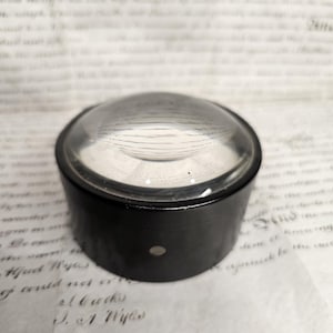 May include: A magnifying glass with a black casing and a clear, curved lens. The glass is resting on a sheet of paper with handwritten script. The lens is domed and reflects the light.