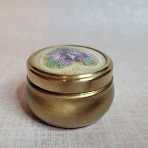 Op de afbeelding: Een klein, rond, goudkleurig metalen doosje met een bloemenmotief op de deksel. De deksel is versierd met een witte achtergrond en een paars en groen bloemenmotief.