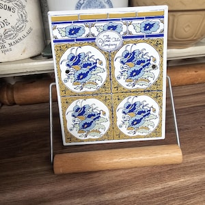 May include: A decorative tile holder with a wooden dowel and metal frame. The tiles feature a blue floral design on a white background, framed by gold and blue borders. The Devon Tile Company logo is visible.