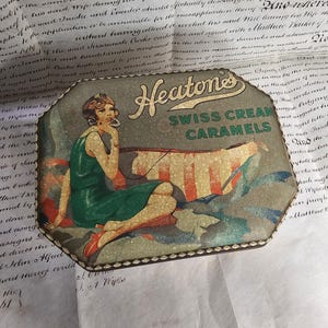 May include: Vintage Heatone's Swiss Cream Caramels tin. The tin features an illustration of a woman in a green dress, red shoes, and a stylish bob haircut. The tin is octagonal and has a decorative border.