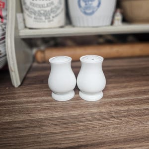 Peut inclure: Une paire de salière et poivrière en céramique blanche avec une silhouette lisse et incurvée en forme de sablier. Chaque pièce présente une base arrondie et un sommet percé de petits trous pour le dosage, formant un ensemble assorti.