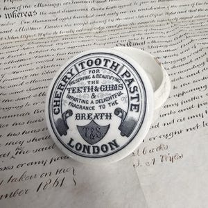 May include: A vintage Cherry Tooth Paste container with a lid. The white ceramic container has a black and gray circular design with text that reads "Cherry Tooth Paste" and "London". The container sits on a background of aged paper.