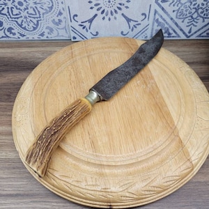 Könnte beinhalten: Ein Schneidebrett aus Holz mit einem geschnitzten Design am Rand. Ein Vintage-Messer mit einem braunen Hirschhorn-Griff liegt auf dem Brett.