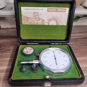 Könnte beinhalten: Ein schwarzes Lederetui mit grünem Innenraum, das ein Tachometer mit silbernem Zifferblatt und schwarzem Zeiger enthält. Das Tachometer hat ein weißes Zifferblatt mit schwarzen Markierungen und roten Zahlen. Das Etui enthält außerdem drei kleine schwarze Gummistücke. Das Etui trägt ein Etikett mit der Aufschrift "Instructions for use Hasler Berne".