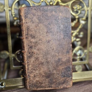 K&ouml;nnte beinhalten: Ein kleines, antikes Buch mit einem abgenutzten, braunen Ledereinband. Der Buchr&uuml;cken ist gen&auml;ht, und die R&auml;nder der Seiten sind sichtbar. Das Buch ist rechteckig und scheint alt zu sein, mit einer verwitterten Textur. Der Hintergrund umfasst einen dekorativen Messingrahmen.