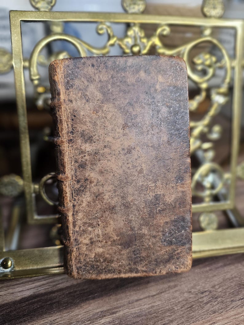 K&ouml;nnte beinhalten: Ein kleines, antikes Buch mit einem abgenutzten, braunen Ledereinband. Der Buchr&uuml;cken ist gen&auml;ht, und die R&auml;nder der Seiten sind sichtbar. Das Buch ist rechteckig und scheint alt zu sein, mit einer verwitterten Textur. Der Hintergrund umfasst einen dekorativen Messingrahmen.
