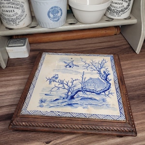 May include: A blue and white ceramic tile with a wood frame. The tile features a blue and white design of a house, trees, and birds. The tile is a decorative piece and would be a great addition to any home.