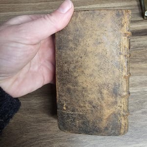 K&ouml;nnte beinhalten: Ein kleines, antikes Buch mit einem abgenutzten, strukturierten Lederumschlag. Der Umschlag ist eine Mischung aus braunen und beigen Farbt&ouml;nen mit sichtbaren Gebrauchsspuren. Der Buchr&uuml;cken hat erhabene B&auml;nder. Das Buch wird in einer Hand gehalten.