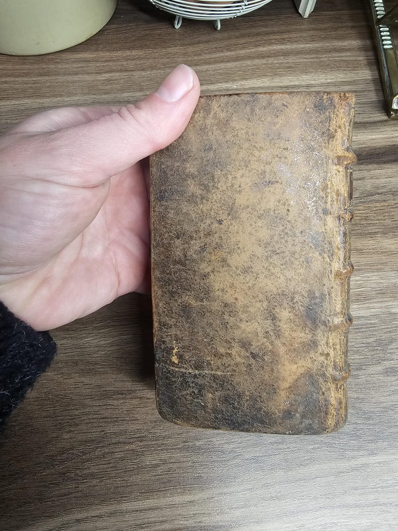 K&ouml;nnte beinhalten: Ein kleines, antikes Buch mit einem abgenutzten, strukturierten Lederumschlag. Der Umschlag ist eine Mischung aus braunen und beigen Farbt&ouml;nen mit sichtbaren Gebrauchsspuren. Der Buchr&uuml;cken hat erhabene B&auml;nder. Das Buch wird in einer Hand gehalten.