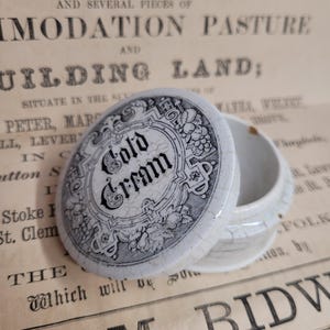 Puede incluir: Un tarro de cerámica blanco con un diseño en blanco y negro. La tapa tiene un diseño en blanco y negro con las palabras "Cold Cream" en una fuente decorativa.