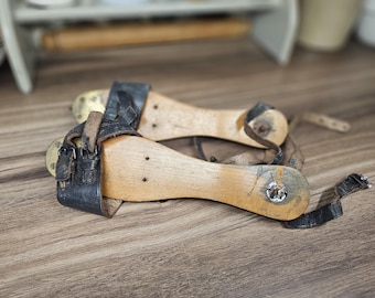 May include: A pair of vintage ice skates featuring wooden soles and black leather straps with metal buckles. The skates have metal blades attached to the underside of the wood, showing signs of wear and oxidation on the metal components and leather.