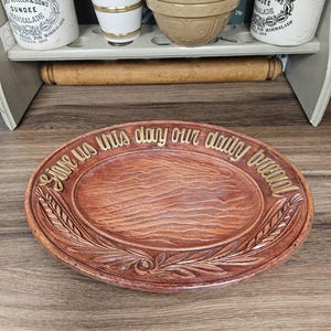 May include: An oval-shaped brown ceramic bread plate featuring a wood grain texture in the centre and wheat stalk motifs along the rim. The plate is embossed with the gold-coloured text "Give us this day our daily bread" along the upper edge.