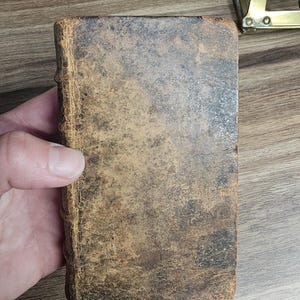 K&ouml;nnte beinhalten: Ein kleines, antikes Buch mit einem abgenutzten, braunen Ledereinband. Der Einband hat ein geflecktes Aussehen mit dunkleren Bereichen und sichtbaren Abnutzungserscheinungen. Der Buchr&uuml;cken ist ebenfalls ledergebunden und weist Altersspuren auf. Das Buch wird in einer Hand gehalten.