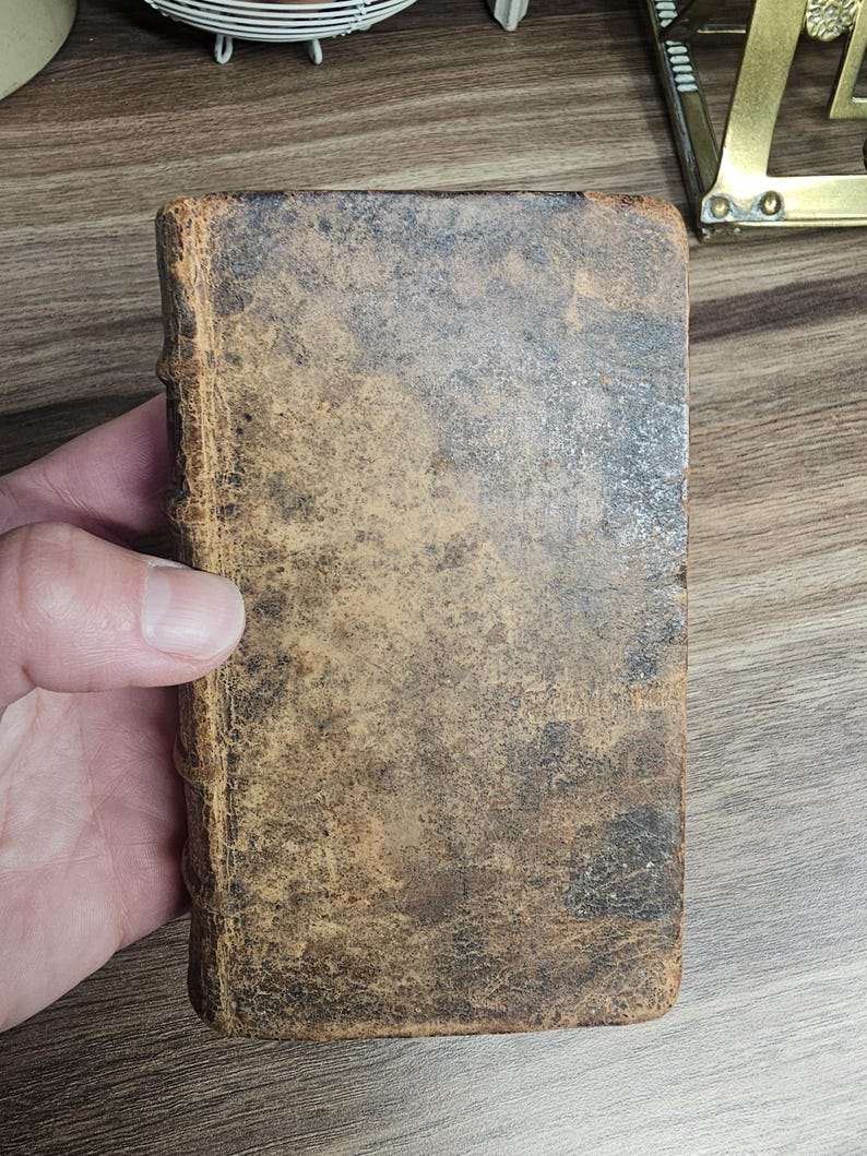 K&ouml;nnte beinhalten: Ein kleines, antikes Buch mit einem abgenutzten, braunen Ledereinband. Der Einband hat ein geflecktes Aussehen mit dunkleren Bereichen und sichtbaren Abnutzungserscheinungen. Der Buchr&uuml;cken ist ebenfalls ledergebunden und weist Altersspuren auf. Das Buch wird in einer Hand gehalten.