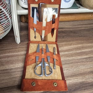 May include: Open brown leather manicure set containing scissors, nail files, and a comb. The tools are held in place by leather straps. The case is secured with two snap closures. The set is open, revealing the tools.