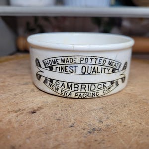 May include: A small, off-white ceramic container with a wide rim. The container has black text that reads "HOME MADE POTTED MEAT FINEST QUALITY CAMBRIDGE NEW ERA PACKING COMPY". The container is on a wooden surface.