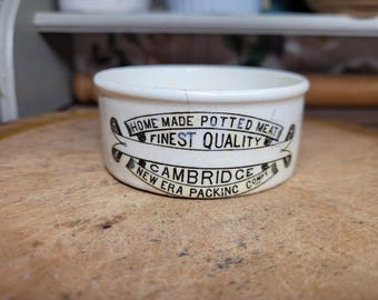 May include: A small, off-white ceramic container with a wide rim. The container has black text that reads "HOME MADE POTTED MEAT FINEST QUALITY CAMBRIDGE NEW ERA PACKING COMPY". The container is on a wooden surface.