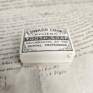 Może przedstawiać: Prostokątny biały ceramiczny pojemnik na mydło do zębów Edward Cooks Hygienic Tooth Soap. Na wieczku znajduje się czarny nadruk z napisem: Edward Cooks Hygienic Tooth Soap, Recommended by the Dental Profession, otoczony ozdobnymi zawijasami.