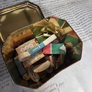 May include: An open, antique gold-colored tin filled with sewing supplies. Contents include spools of thread, buttons, ribbon, and small packages. The tin sits on a vintage document with handwritten text.