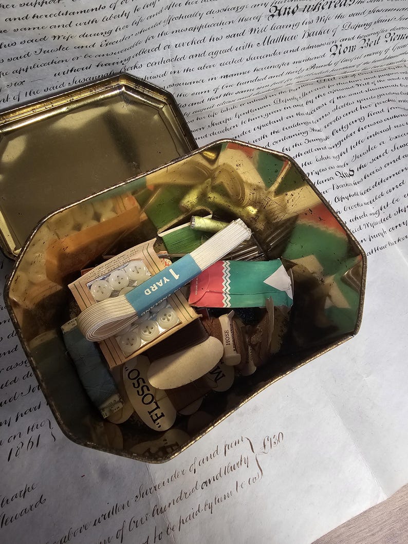 May include: An open, antique gold-colored tin filled with sewing supplies. Contents include spools of thread, buttons, ribbon, and small packages. The tin sits on a vintage document with handwritten text.