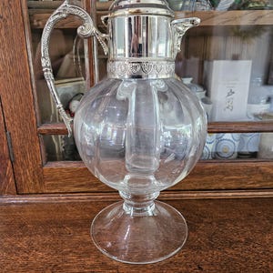 May include: A clear glass claret jug featuring a silver-plated metal collar, a hinged lid with a small finial, and an ornate, curved handle. The vessel has a bulbous, ribbed body and a wide, circular glass base, showcasing a classic design.