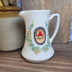May include: White ceramic pitcher with a handle, featuring a circular design with the text "Bass & Co's Pale Ale" and a red triangle with the word "Bass" inside. The design is surrounded by green leaves and yellow hops.