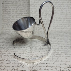 May include: An antique silver egg-shaped cup with a handle, on a stand. The cup is made of polished metal and has a shiny finish.
