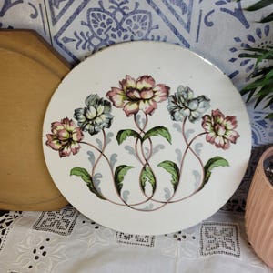 May include: A white ceramic plate with a floral design. The plate features five pink, white, and blue flowers with green leaves and stems. The flowers are arranged in a symmetrical pattern.