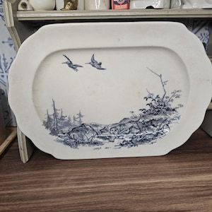 May include: An oval ceramic serving platter with a cream-colored base and a blue transferware print. The design features a landscape with a dog in the foreground, trees, and two birds in flight. The platter has a scalloped edge and a smooth finish.