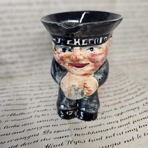 May include: A small, vintage ceramic figurine of a sailor with the word "CHEERIO" on his hat. The figurine is painted with a black hat, navy blue uniform, and a smiling face. The figurine is holding his hands together.
