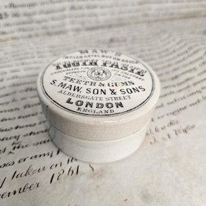 May include: A round, off-white ceramic pot with a lid featuring black printed text that reads: Maw's Indian Betel Nut or Areca Tooth Paste, Unequalled for its salutary effects on the teeth & gums, S. Maw, Son & Sons, Aldersgate Street, London, England.