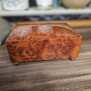 Puede incluir: Una caja rectangular de madera con un acabado pulido y un patrón de veta de madera nudosa prominente. La caja tiene una tapa con bisagras y se apoya sobre cuatro pequeñas patas redondeadas en la base, mostrando un color marrón cálido y rico.