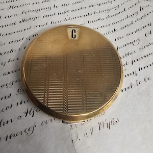 Op de afbeelding: Een ronde, goudkleurige compactpoederdoos met een gestructureerd, geometrisch patroon. De compactpoederdoos heeft een klein, rechthoekig accent met de letter "C" in zwart. De compactpoederdoos ligt op verouderd papier met handschrift.