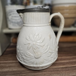 以下が含まれることがあります： 白い陶器のピッチャーで、デイジーの花柄のデザインです。ピッチャーには取っ手と金属製の蓋が付いています。ピッチャーは木製の表面に置かれています。ピッチャーの高さは約18cmです。