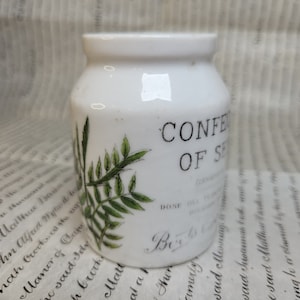 Puede incluir: Tarro de cerámica blanco con un diseño de helecho verde y el texto "CONFECTIONS OF SENNA" impreso en la parte delantera. El tarro tiene una tapa y probablemente se usa para almacenar hierbas medicinales.