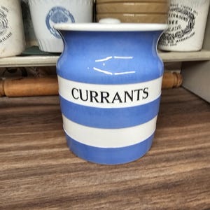 May include: A blue and white ceramic jar with the word "CURRANTS" in black lettering. The jar has a lid and is decorated with horizontal blue and white stripes. Other jars are visible in the background.