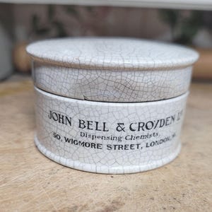 Może przedstawiać: Antyczny, kremowy ceramiczny słoik apteczny z efektem spękanego szkliwa. Cylindryczny słoik ma pokrywkę i zawiera napis "JOHN BELL & CROYDEN" oraz "Dispensing Chemists, 50, Wigmore Street, London W".