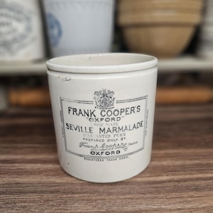 May include: A vintage white ceramic jar with the text "FRANK COOPER'S OXFORD SEVILLE MARMALADE" in black. The jar has a cylindrical shape and sits on a wooden surface. Other ceramic containers are in the background.